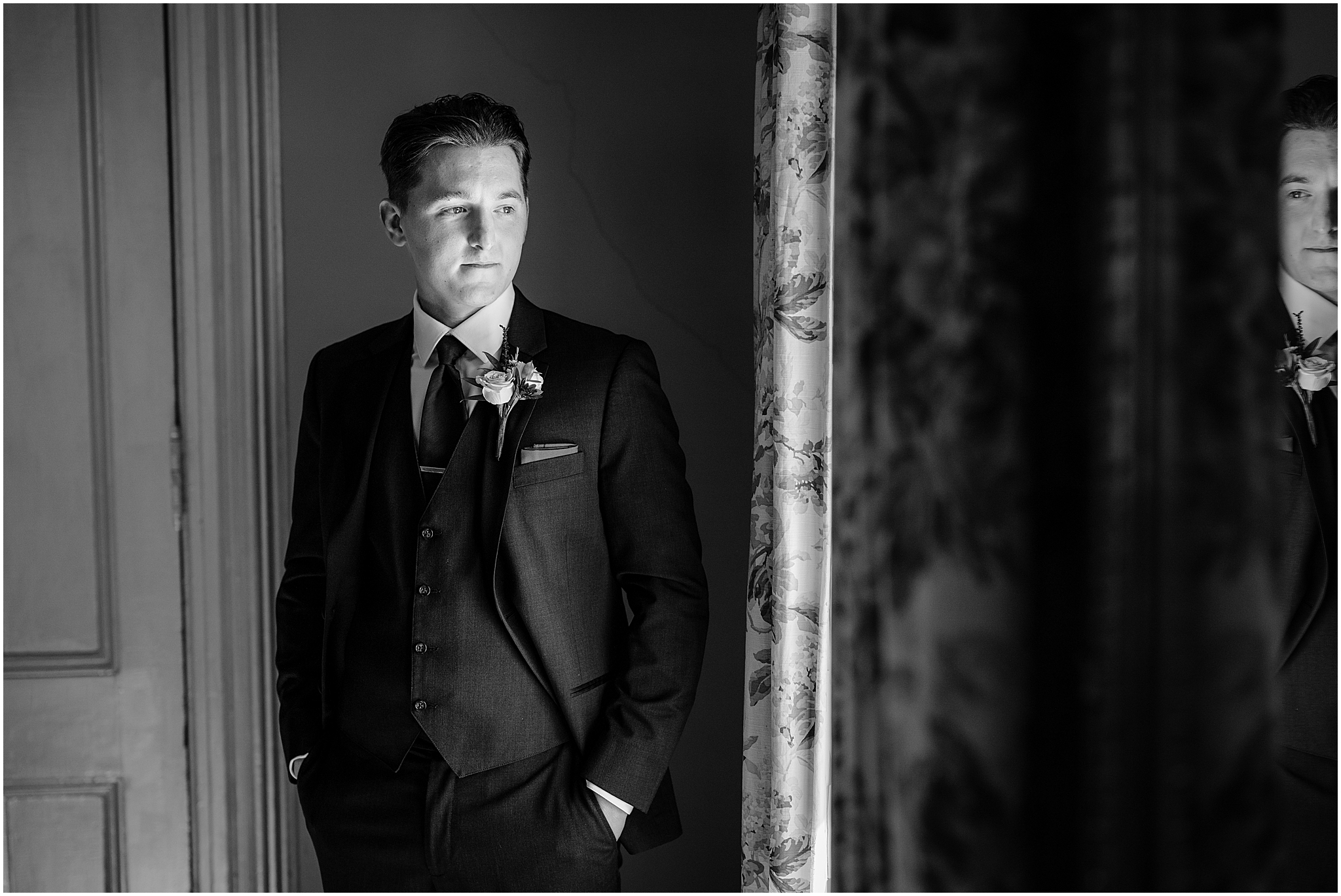 groom stands with hands in pocket of dark suit looking out the window at the Feast at Round Hill