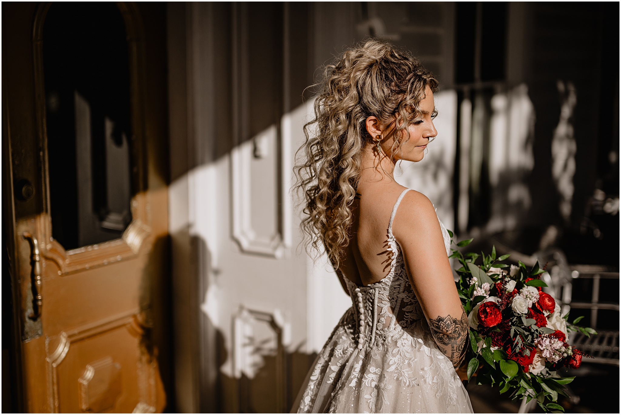 blonde bride with curls in hair looks over shoulder in wedding dress with corset bodice while holding red and white bouquet 