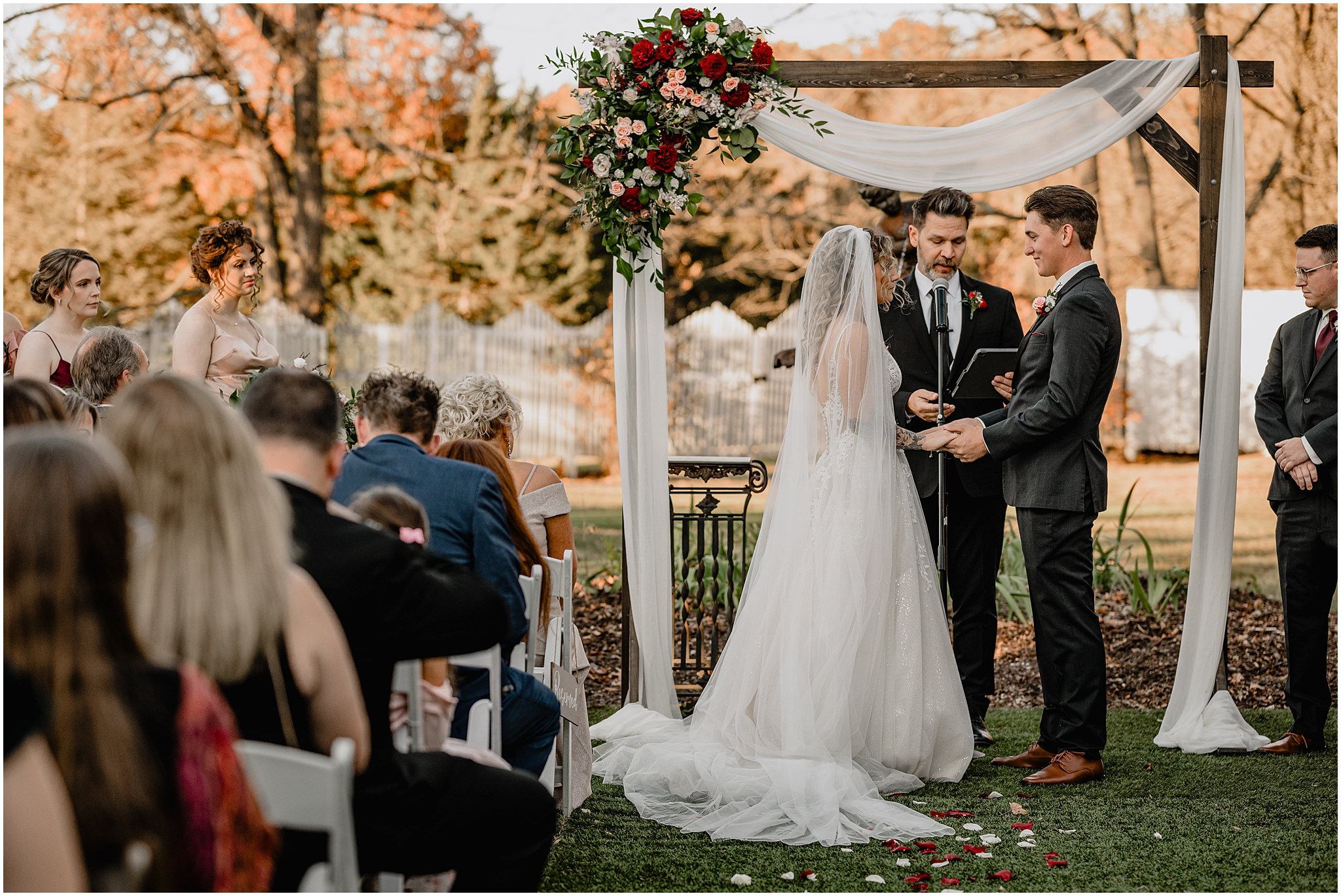 bride and groom hold hands under wooden arbor with spray of pink and red roses on top left during ceremony at the Feast at Round Hill