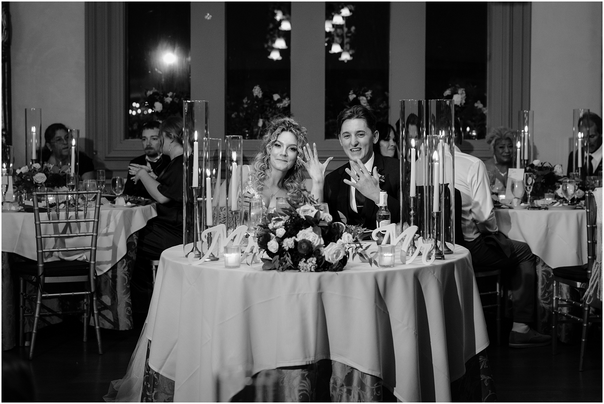 bride and groom hold up hands with wedding bands sitting at sweetheart table in center of the room during wedding reception at the Feast at Round Hill