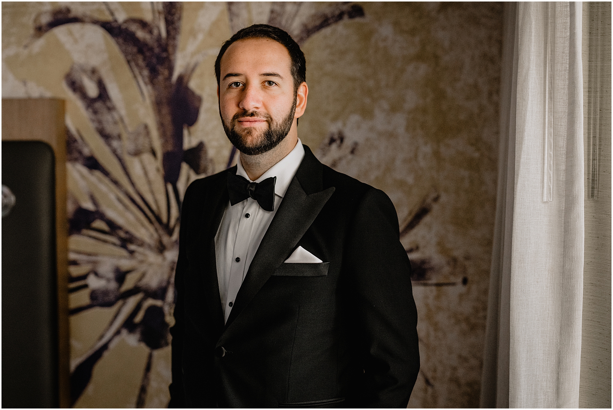 groom stands in black tux in front of the wall in suite at Tappan Hill Mansion