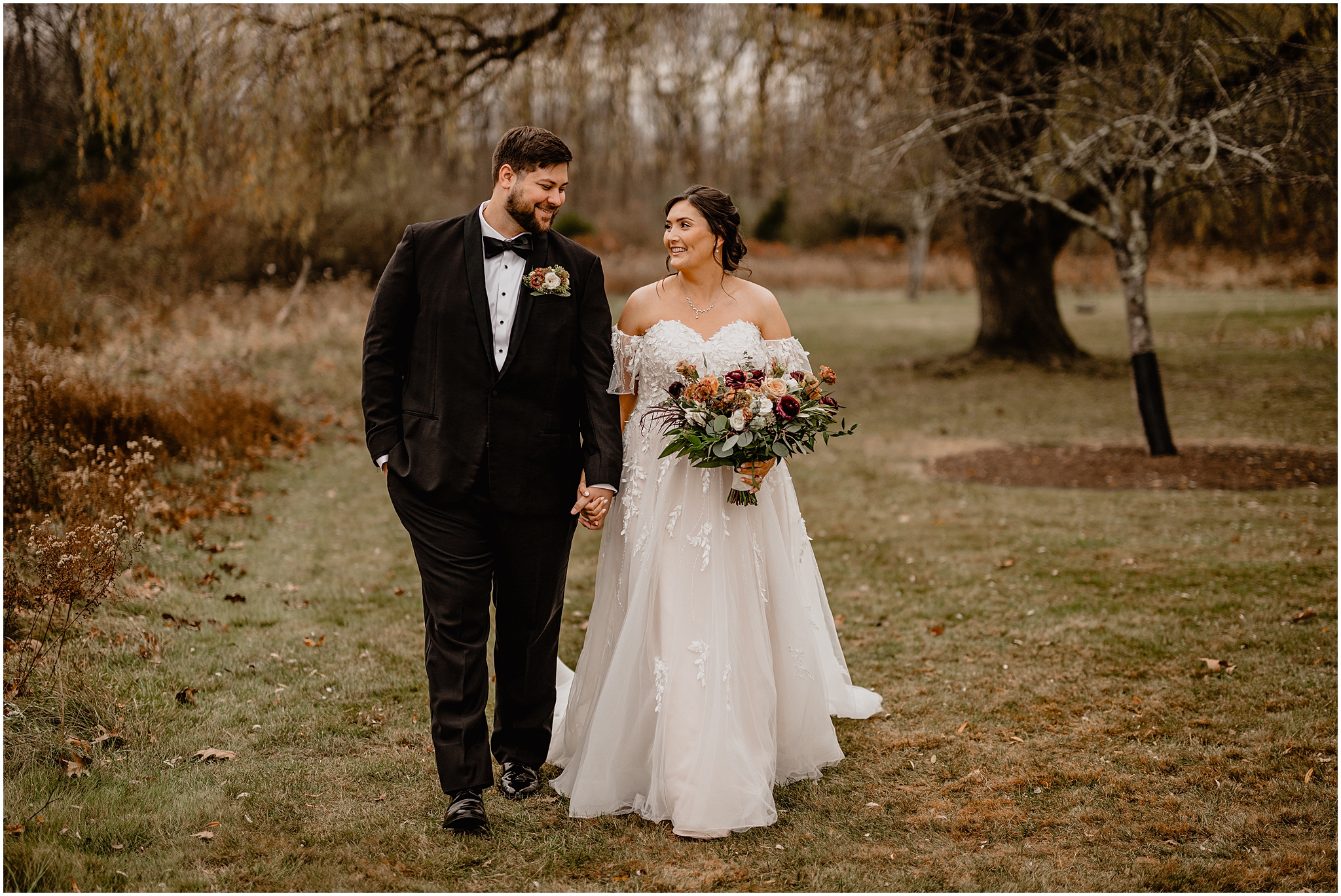 bride and groom hold hands walking through field while bride holds bouquet of fall flowers 