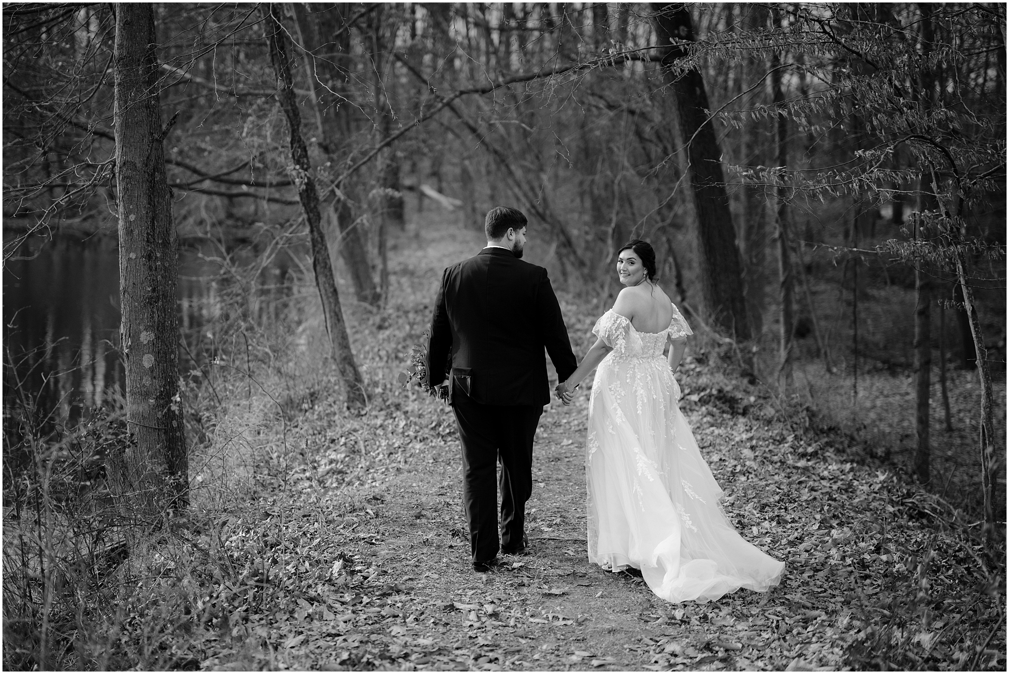 bride and groom hold hands walking through woods with bride looking over shoulder 