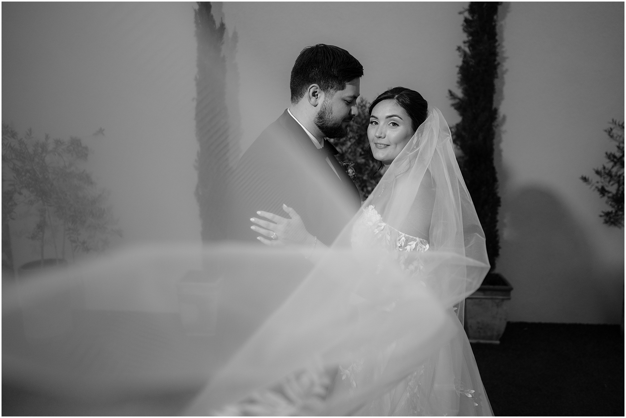 groom smiles at bride while she looks towards the camera with the veil pulled in front of them