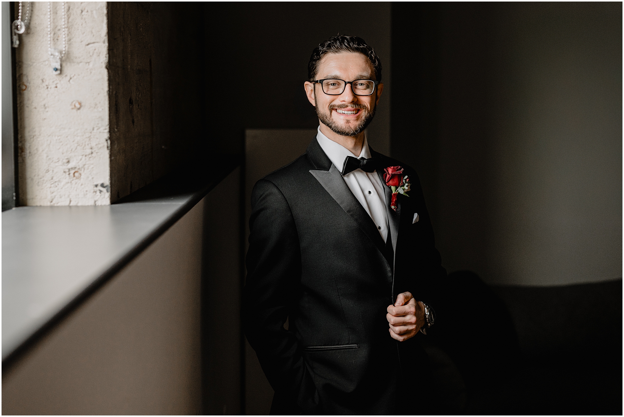 groom stands with hand on lapel of black suit with red rose inside Philadelphia hotel
