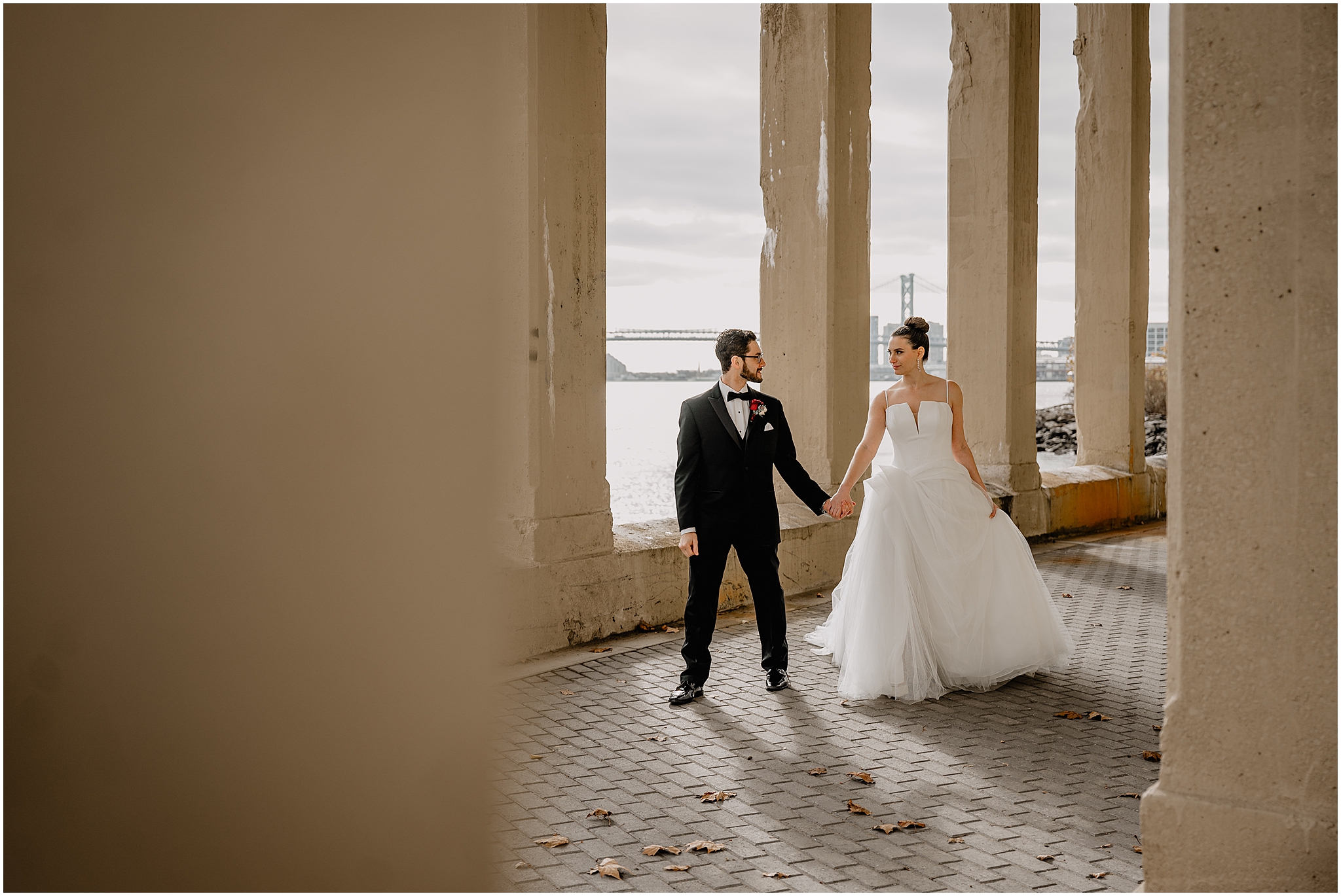 newlyweds hold hands under bridge at Penn Landing along the Delaware River