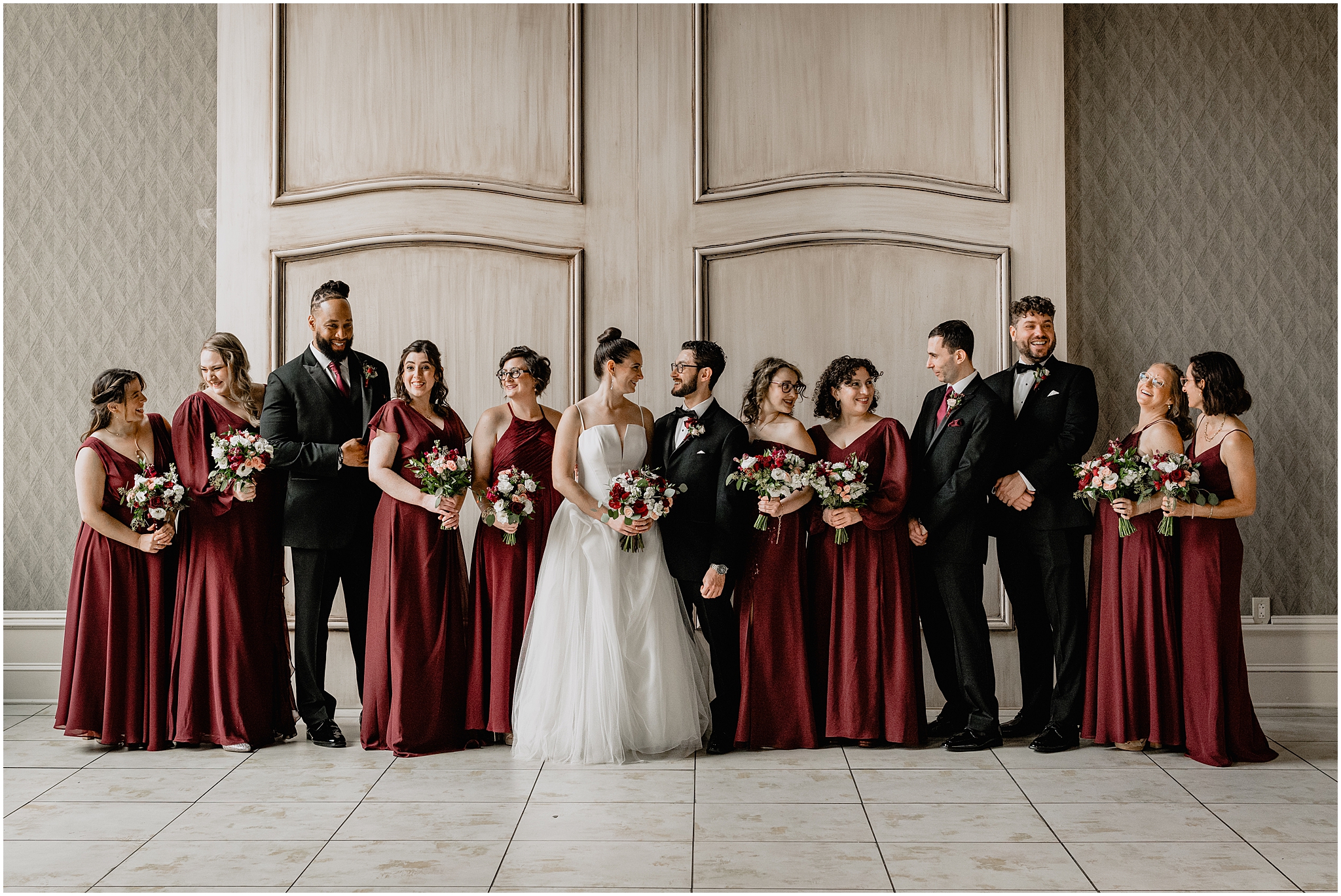 newlyweds stand with wedding party in burgundy bridesmaid gowns and black suits