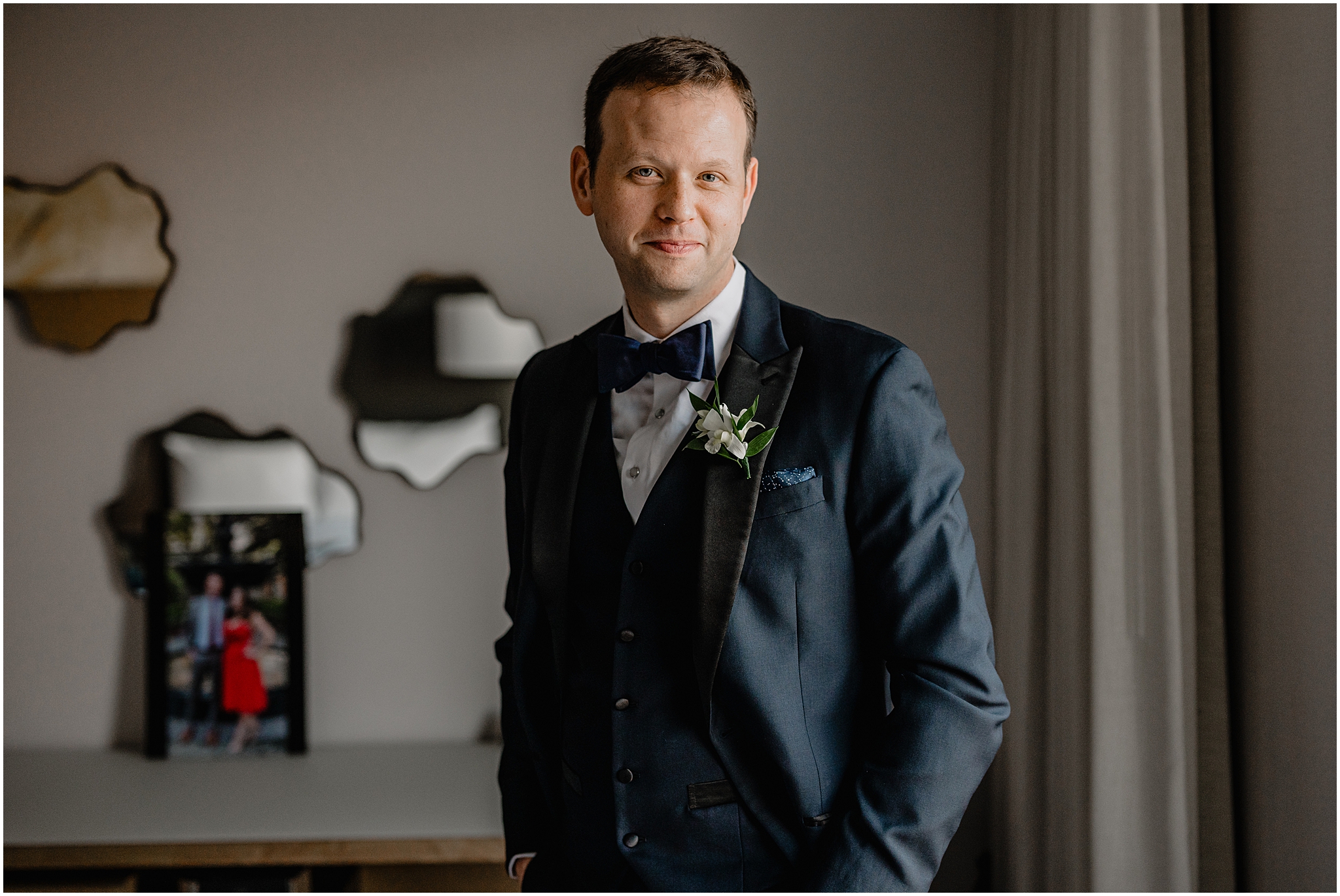 groom in black suit stands with hands in pockets inside Hudson Valley hotel room