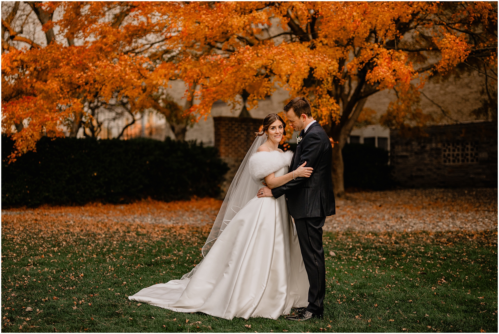 groom looks down at bride in wedding gown with white fur in front of trees with orange leaves in the Hudson Valley