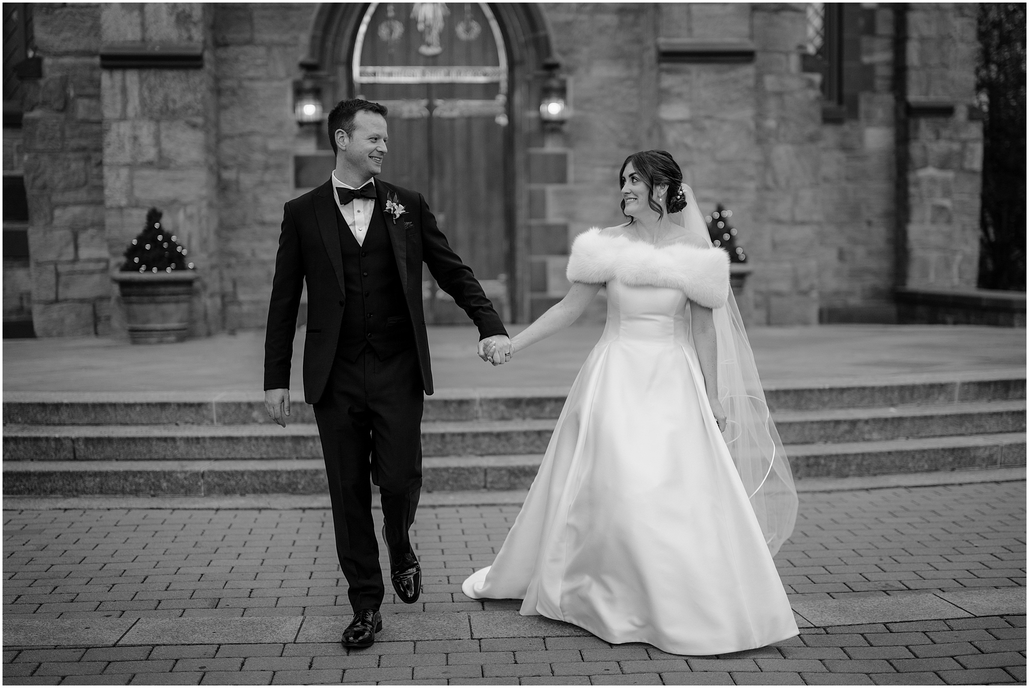 bride in wedding dress with white fur walks with groom in front of stone church in the Hudson Valley