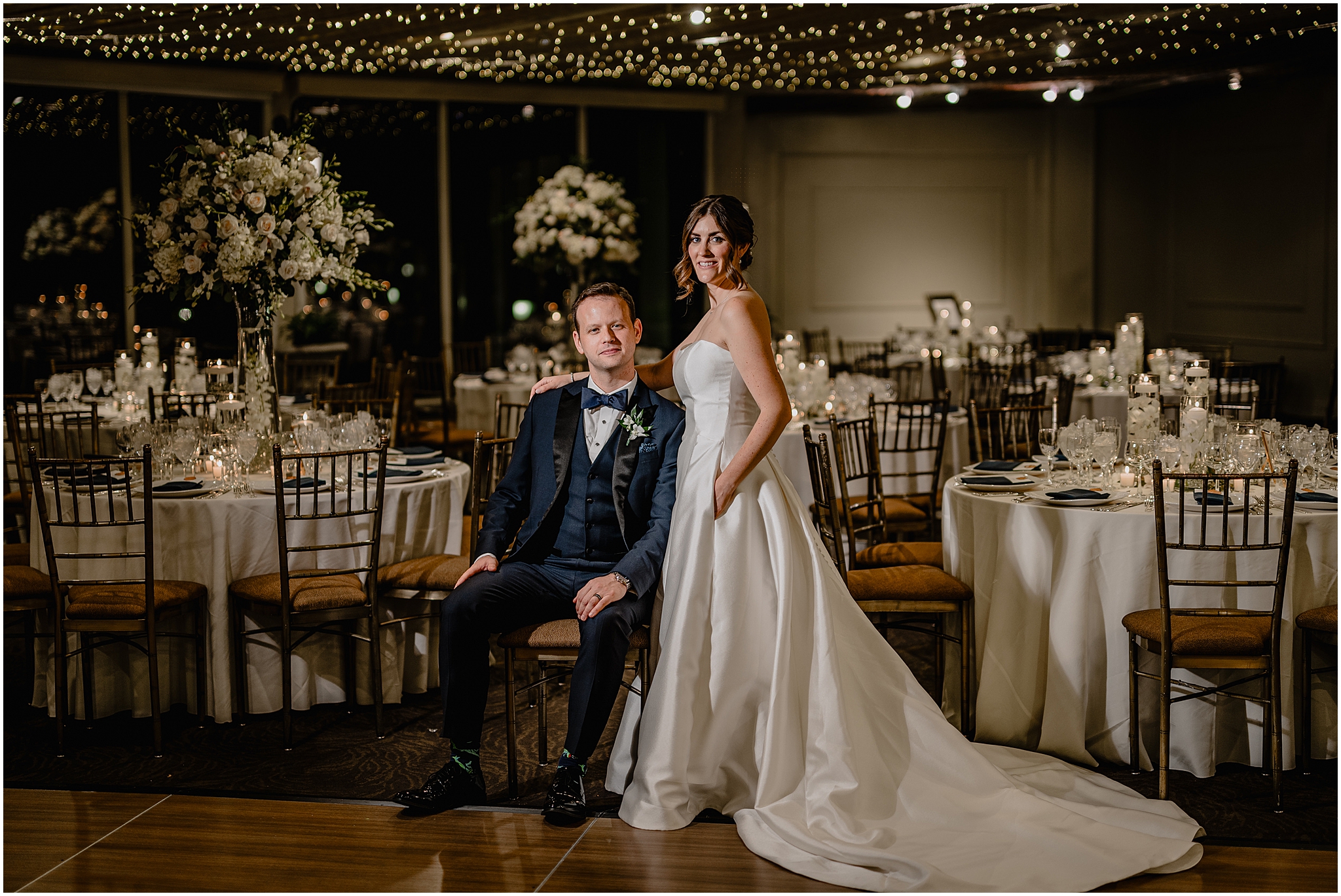 groom sits in chair with bride standing next to him under string of lights inside ballroom at Tappan Hill Mansion