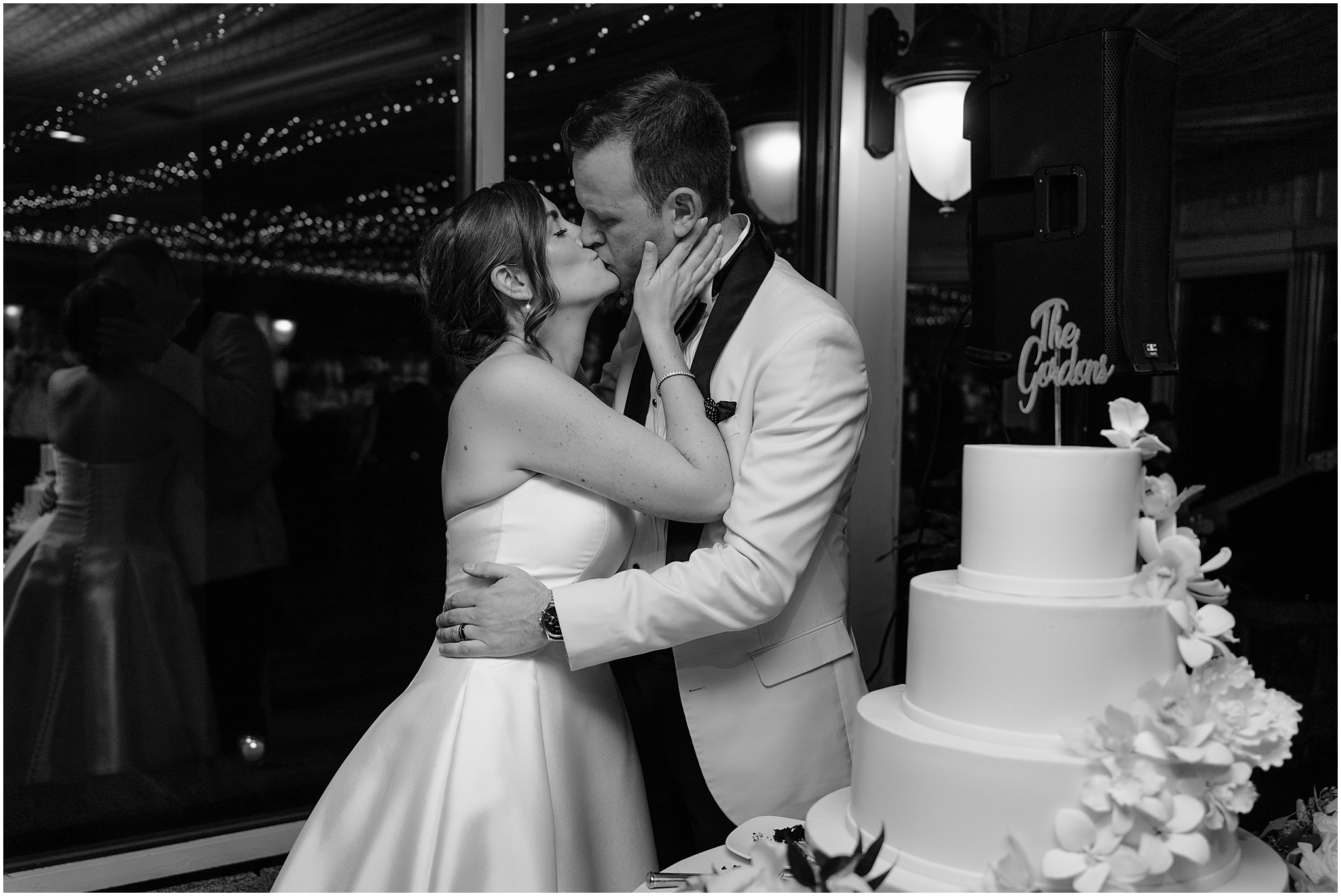 bride and groom kiss by tiered wedding cake with white floral accents at Tarrytown NY wedding reception