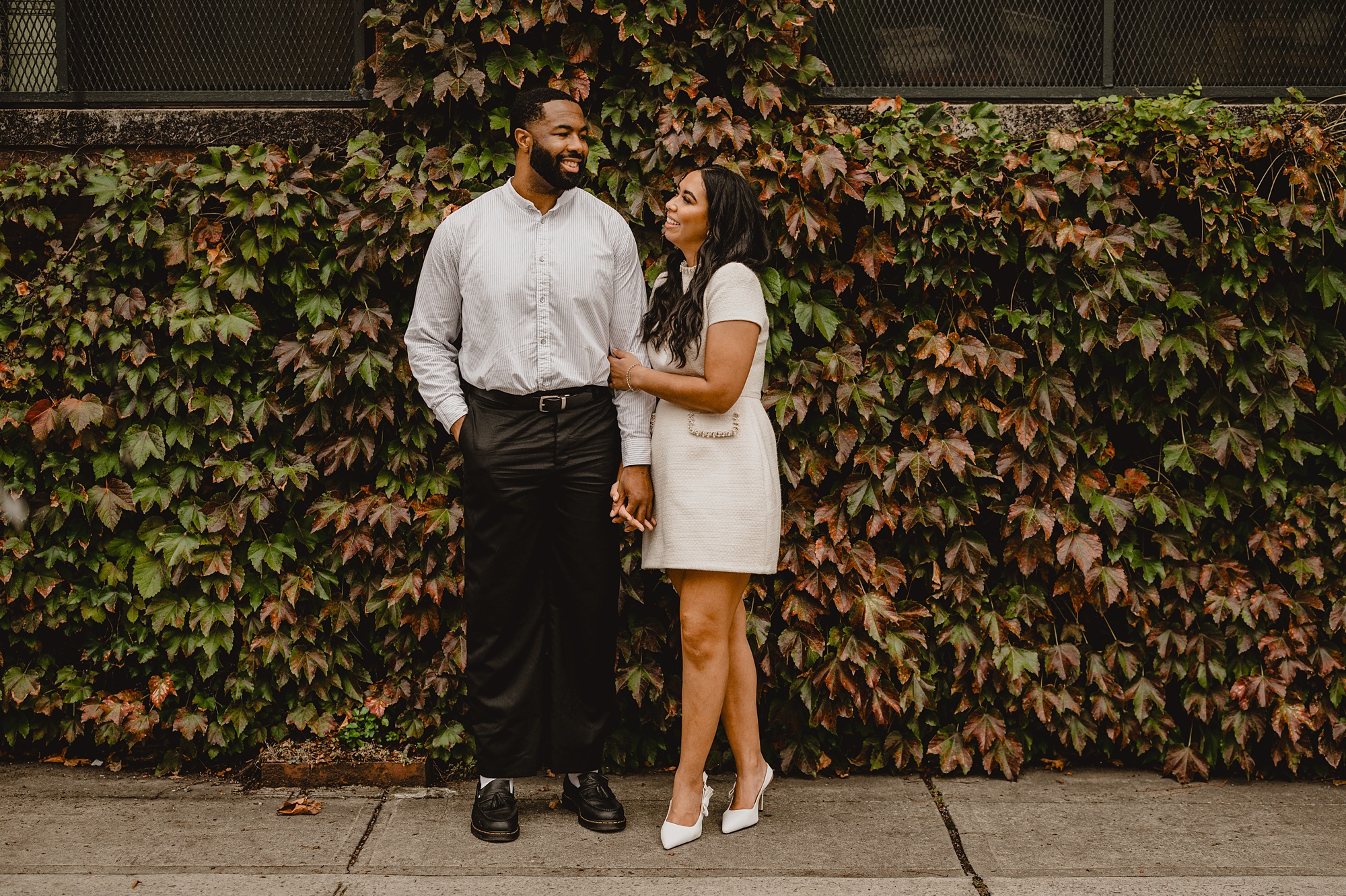 bride holds groom's arm leaning into him outside ivy covered wall at The Foundry on their Long Island Engagement Session