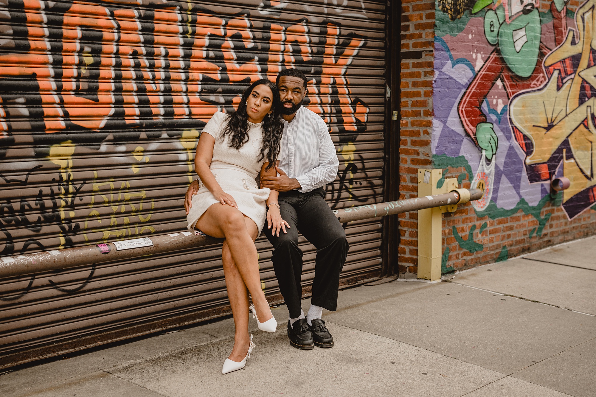 couple sits on metal railing in front of graffiti mural on building for their Long Island Engagement Session