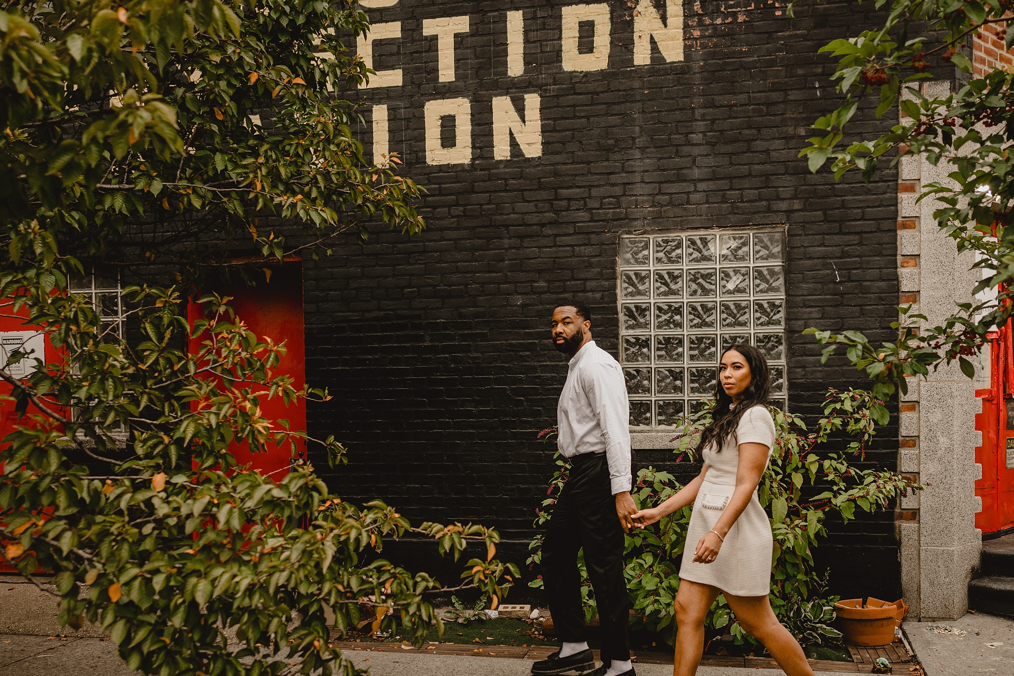 couple holds hands walking by garden and black brick wall in Long Island City