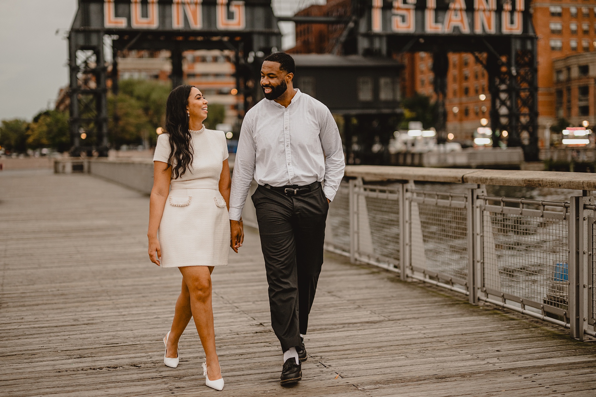 couple walks on dock in front of Long Island City buildings at Gantry State Park Plaza