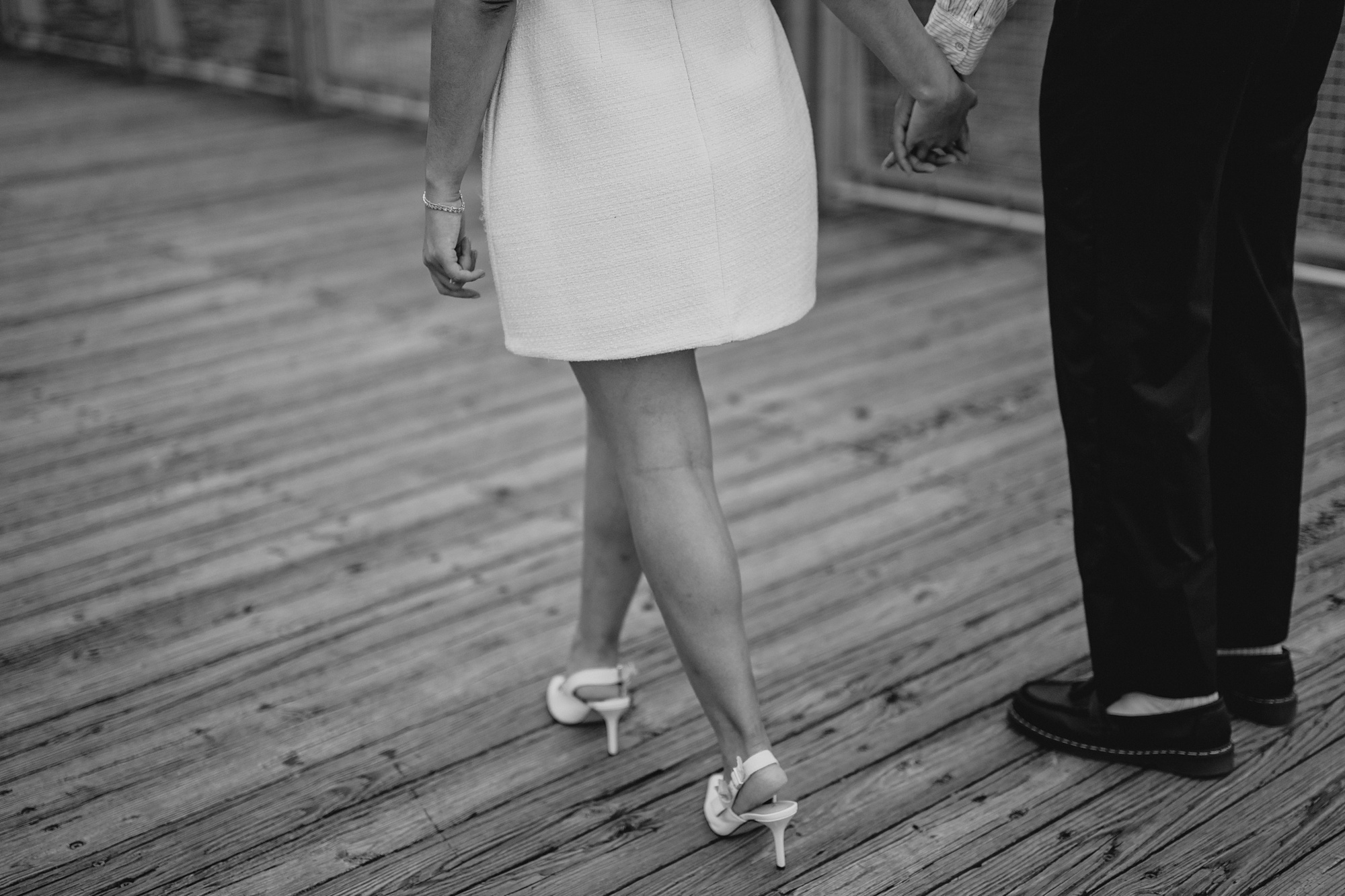woman in white dress and heels holds hands walking with man on wooden dock