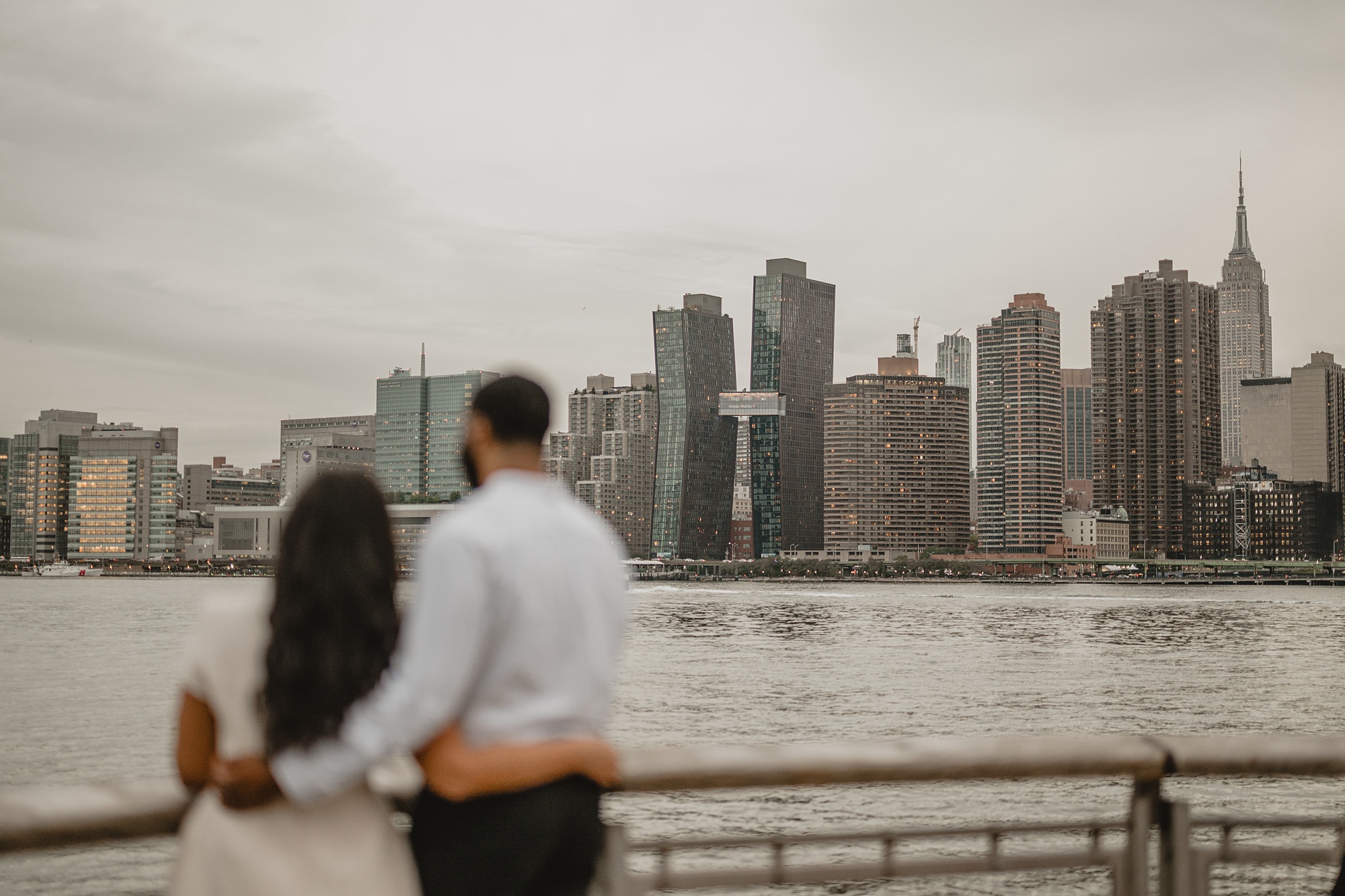 man hugs women in front of railing overlooking the water and NYC skyline from Gantry State Park Plaza