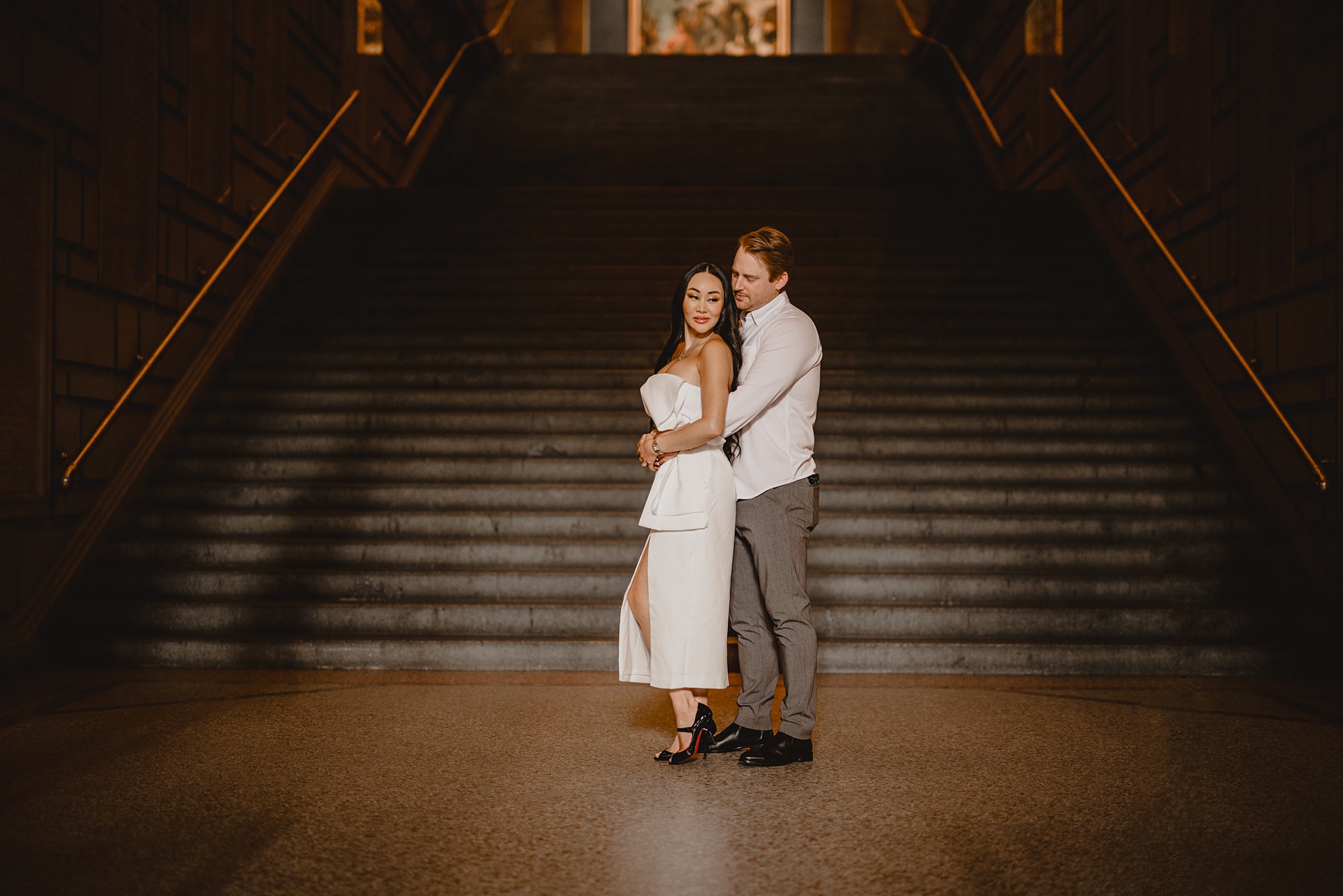 man hugs woman in chic white dress in front of staircase inside the Met Museum