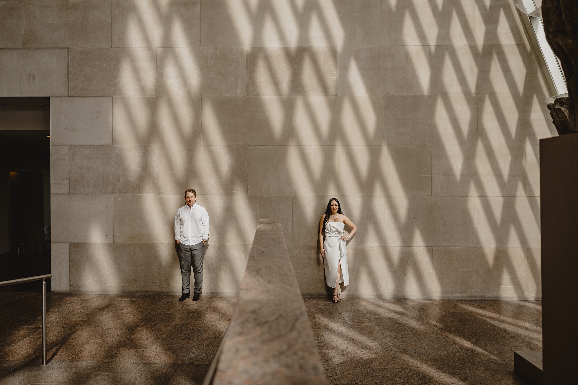 man and woman lean against light tan wall in shadow of windows