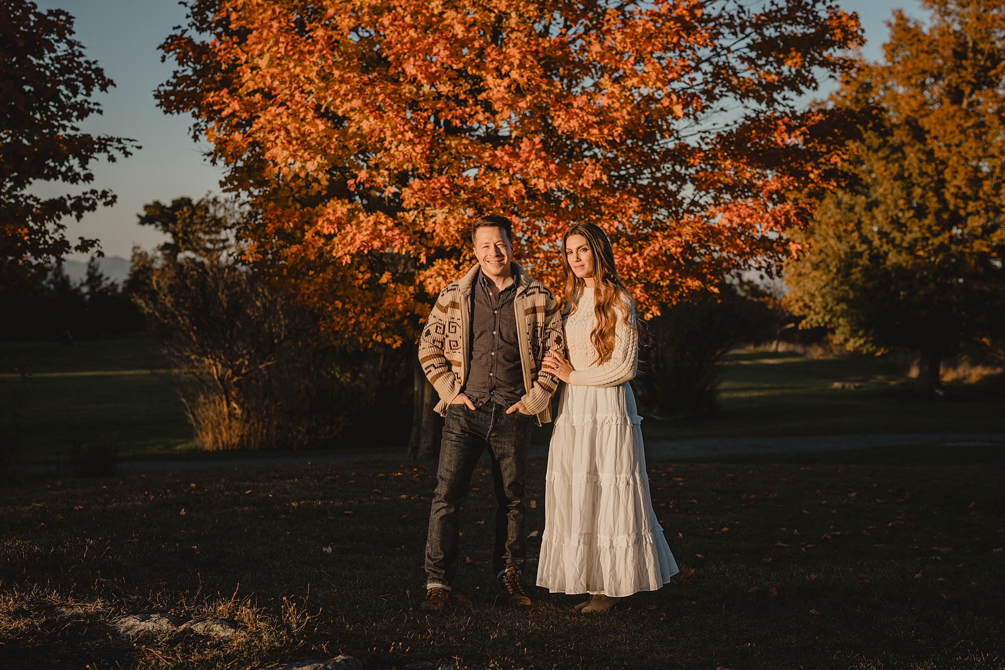woman in white dress talks with man in cozy sweater in front of tree with orange leaves