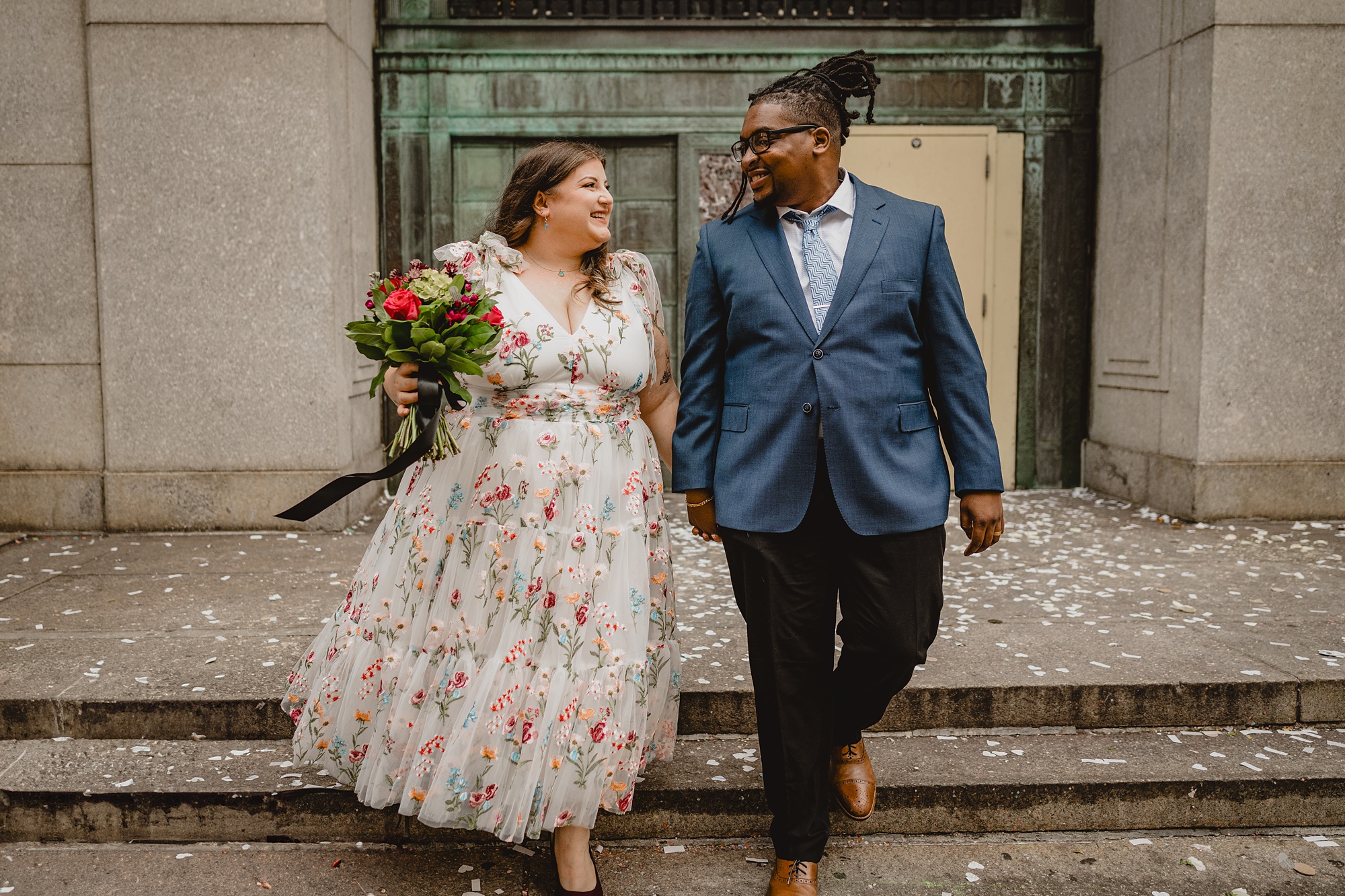 bride in floral dress holds hands with groom in blue suit leaving NYC City Hall
