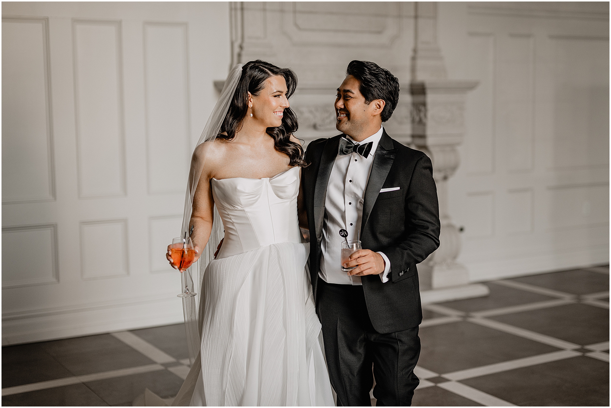 bride holds drink smiling at groom in front of fireplace at the Estate at Florentine Gardens