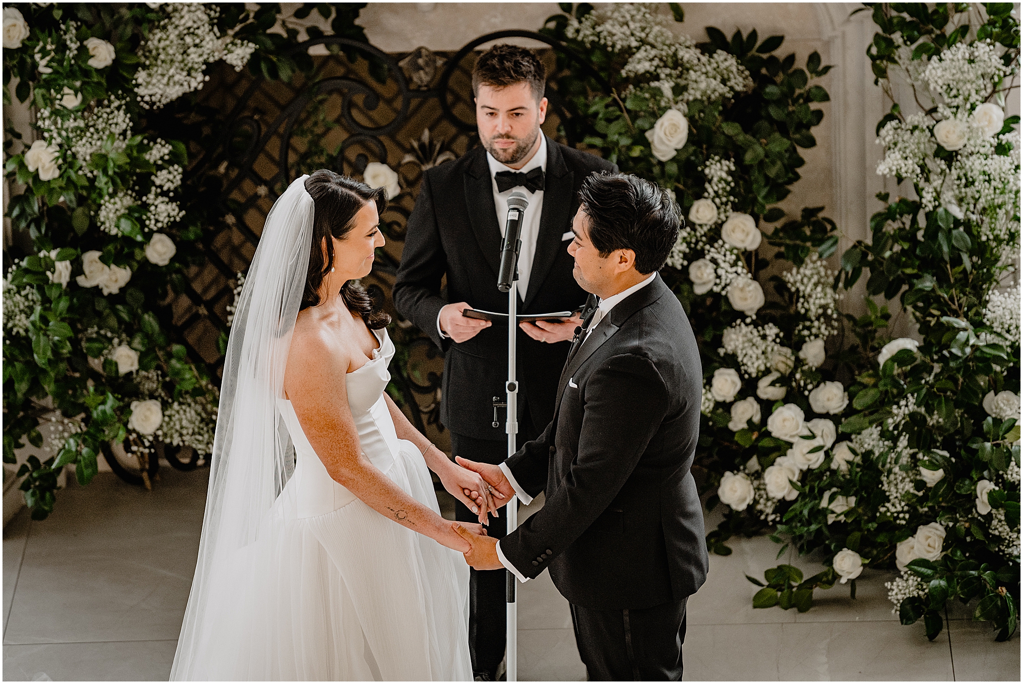newlyweds hold hands listening to officiant in front of white flower sprays along fireplace 