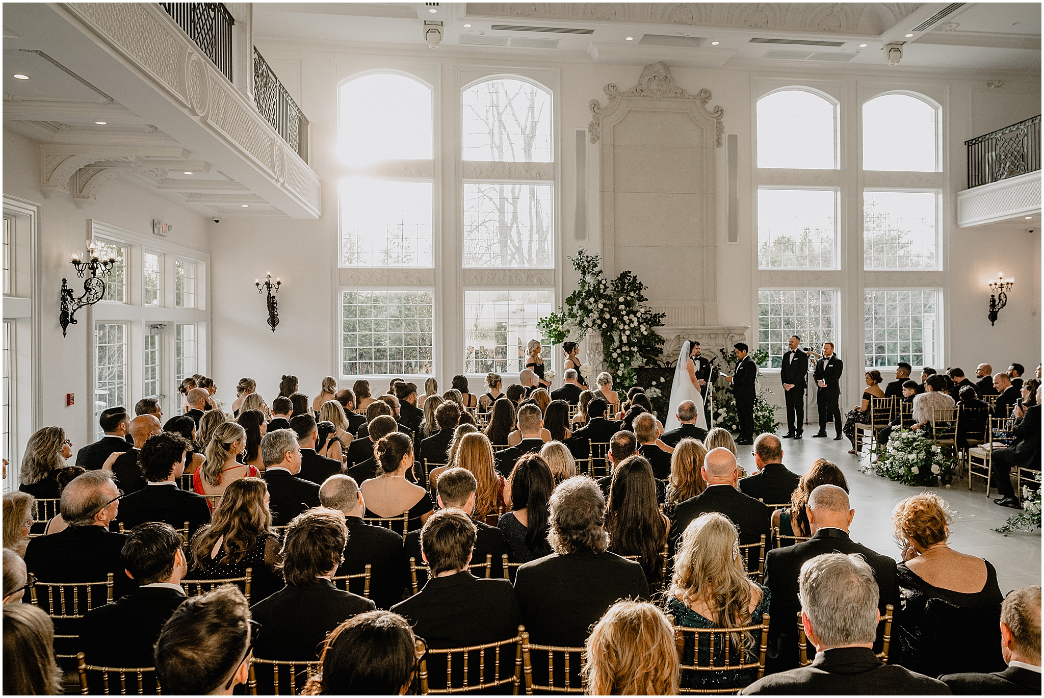 newlyweds hold hands during wedding ceremony in front of fireplace inside the Estate at Florentine Gardens