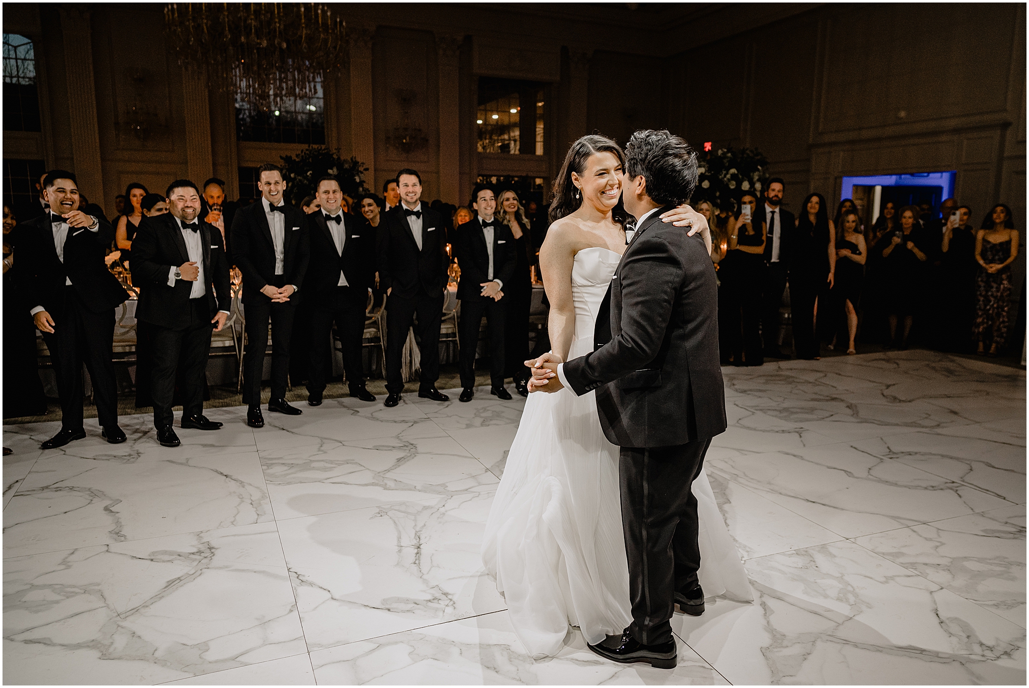 bride and groom have first dance on marbled dance floor surrounded by wedding party 