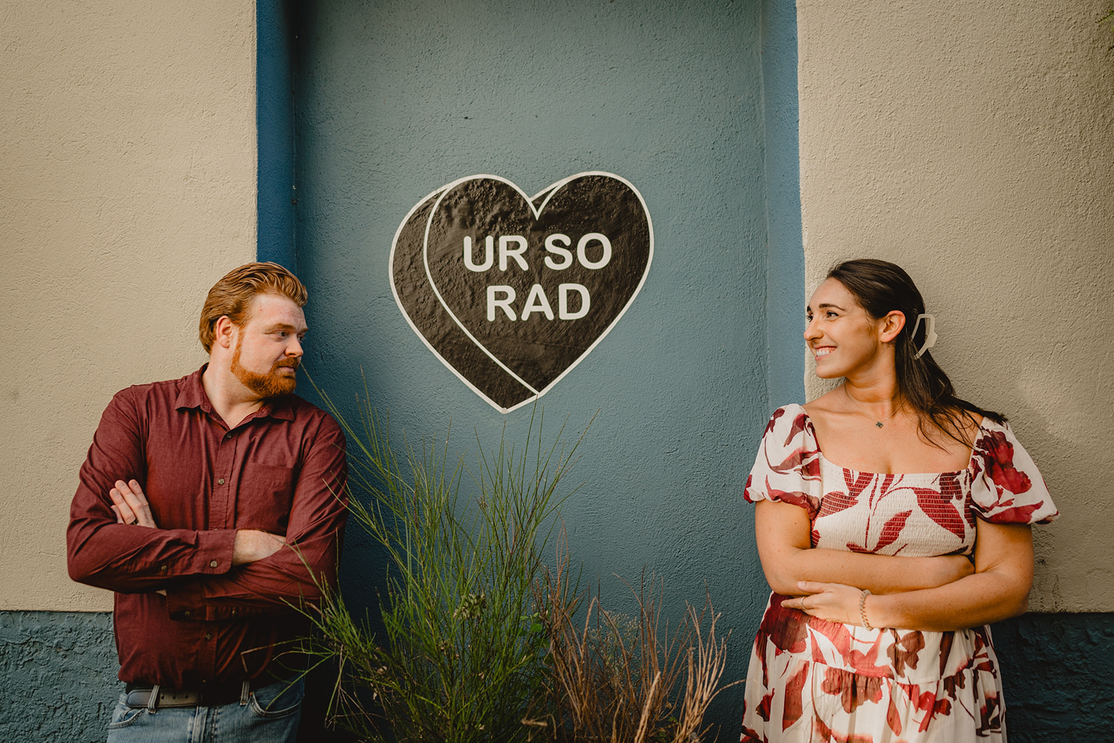 couple smiles standing on either side of black heart that says UR SO RAD