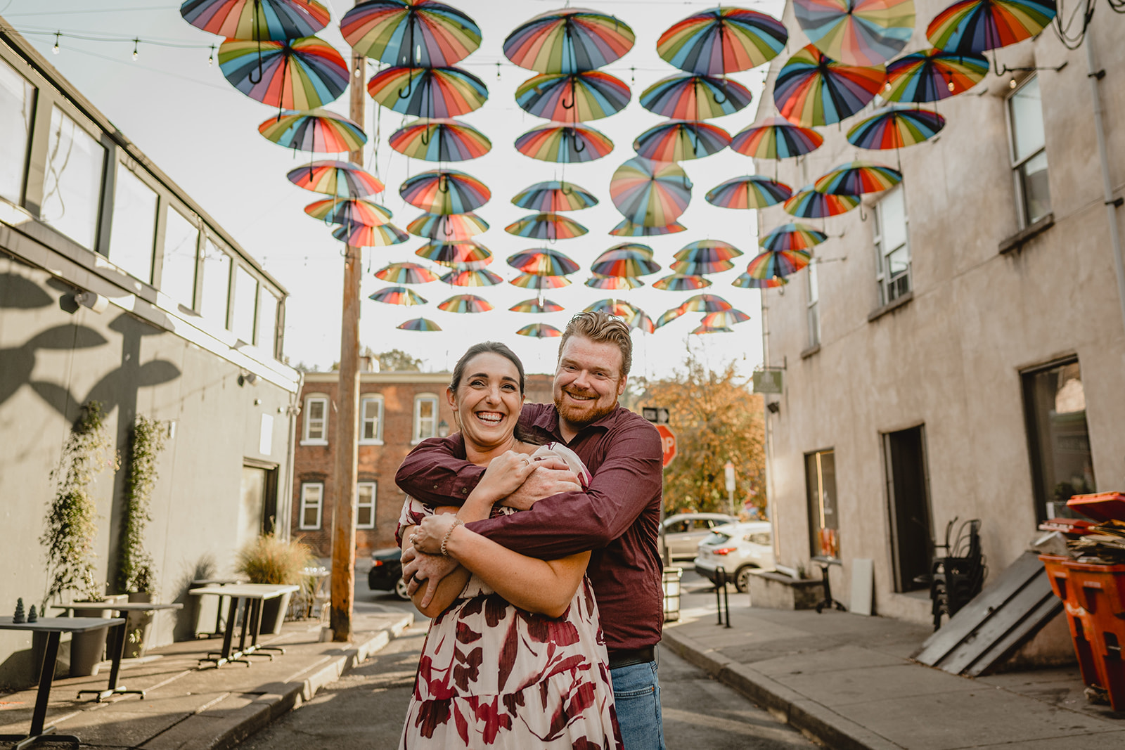 man hugs woman from behind on street under umbrellas during Manayunk engagement session in Philly PA