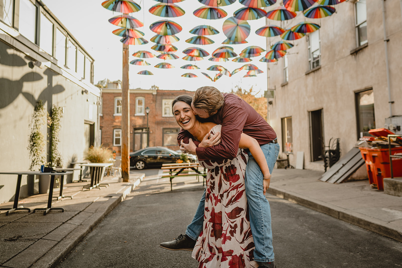 man kisses woman during piggy back ride under hanging umbrellas during Manayunk engagement session in Philly PA