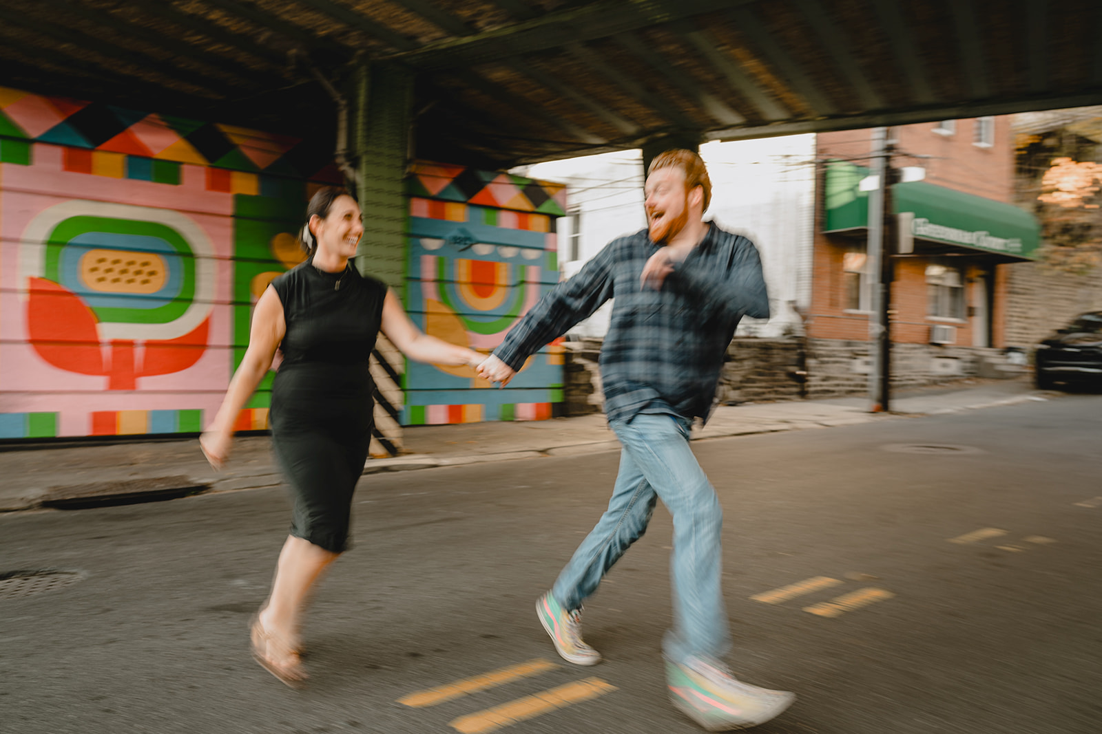 man leads woman across street laughing during Manayunk engagement session in Philly PA