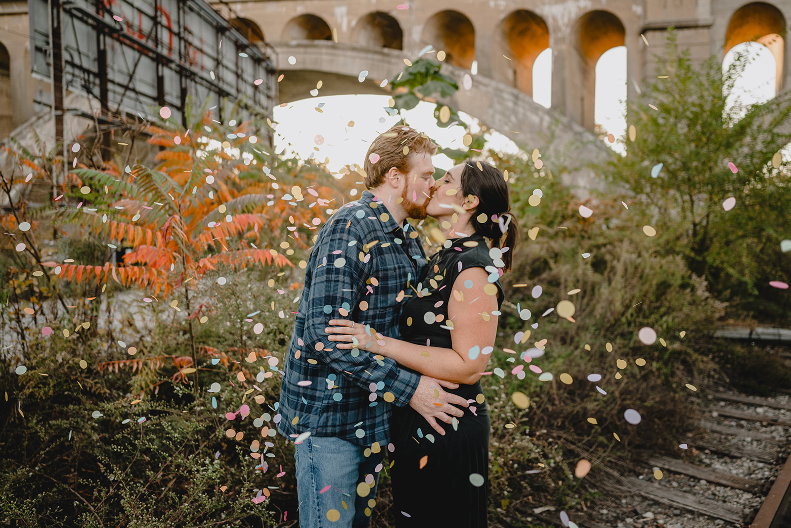 engaged couple kisses by bridge and buildings while confetti floats around them in Manayunk neighborhood