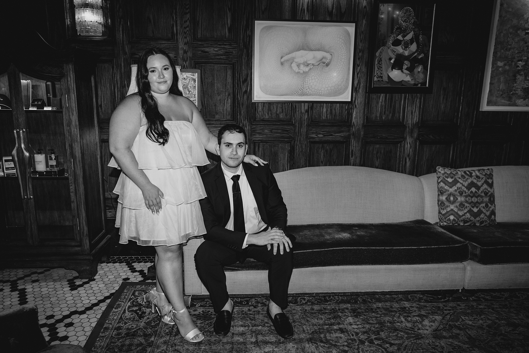 man sits on couch and woman stands next to him in white dress hugging his shoulder 