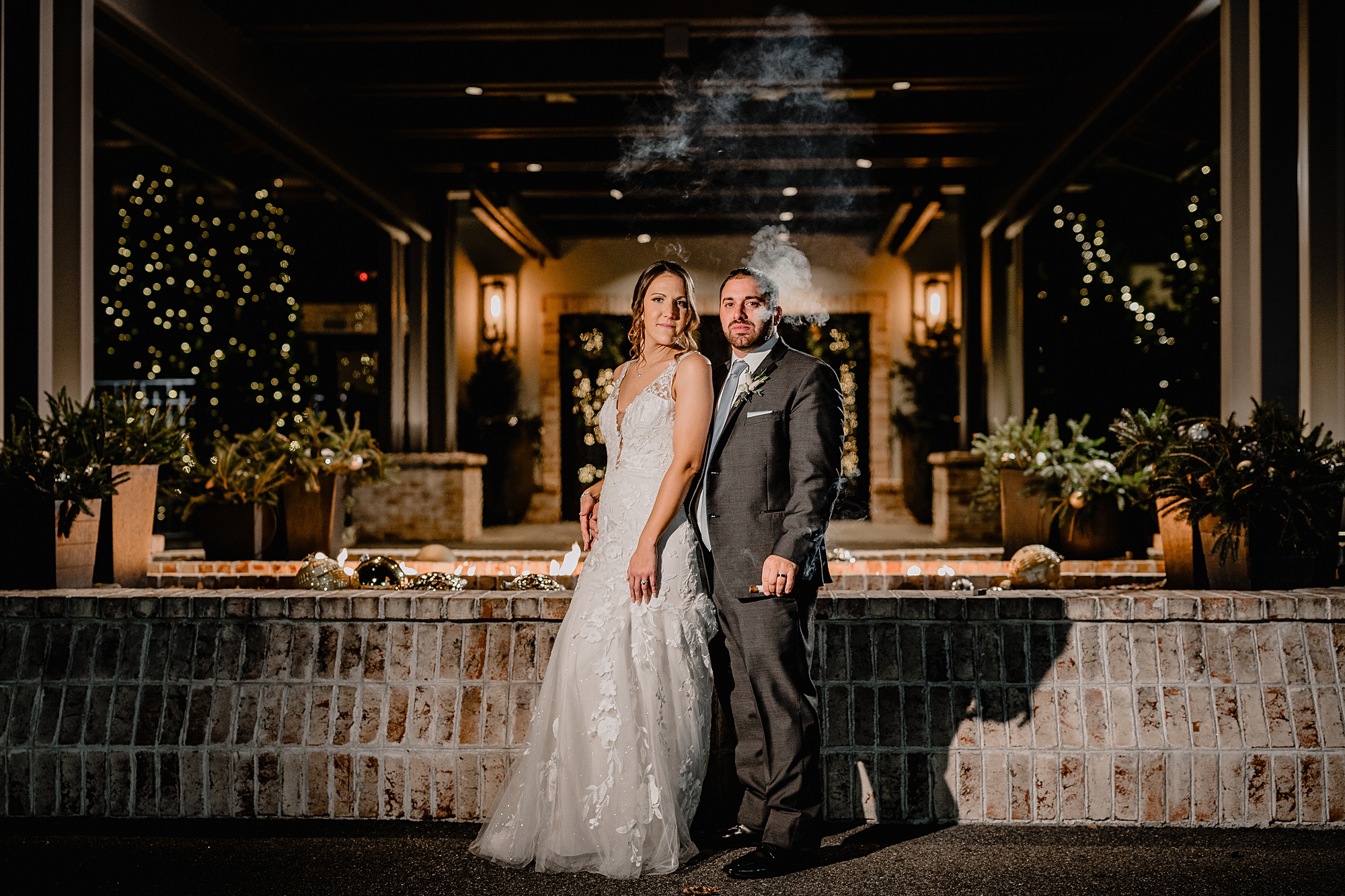 couple poses in front of bar with groom smoking cigar at the Refinery at Perona Farms