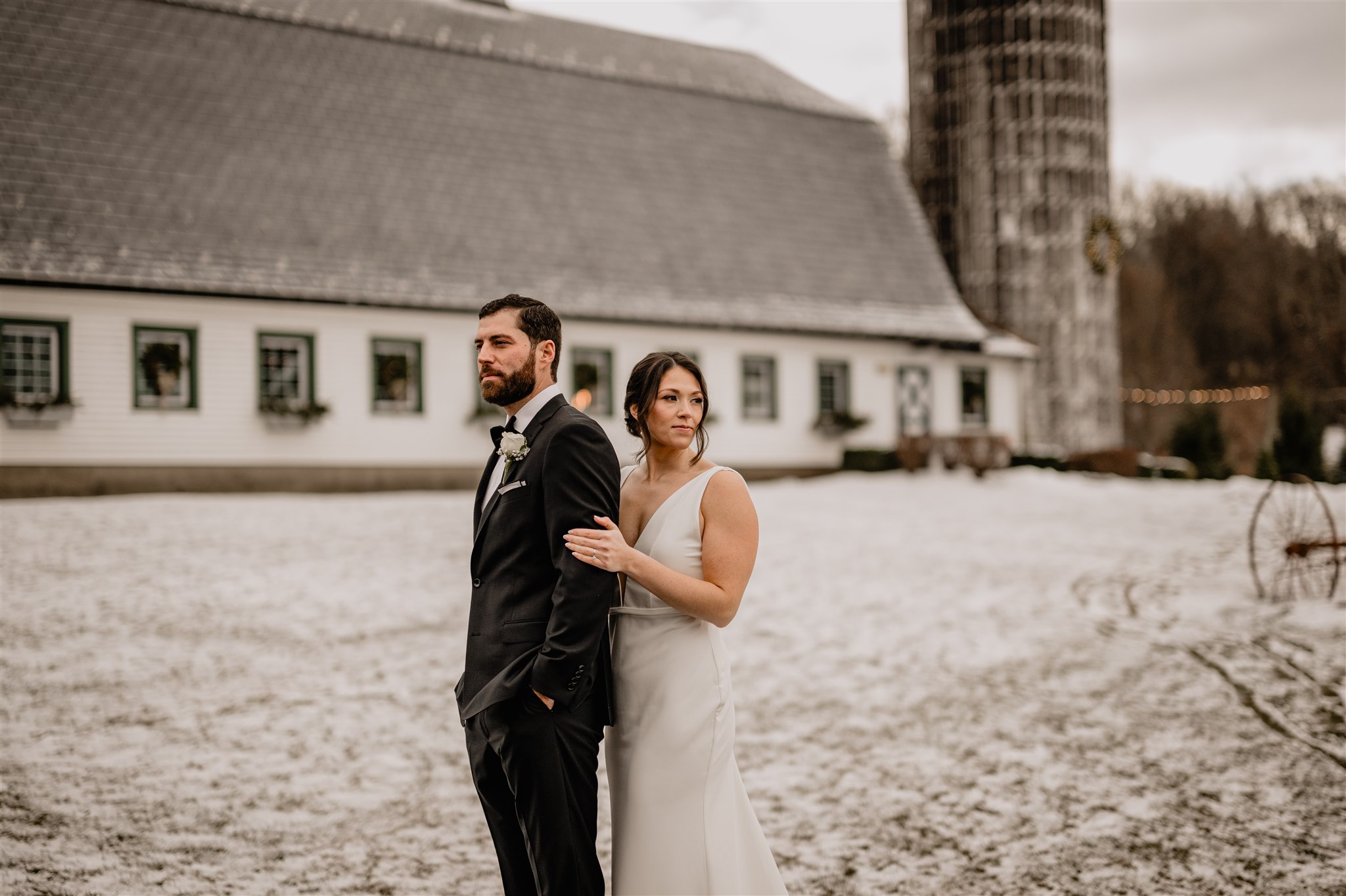 bride stands behind groom holding his arm in front of the Barn at Perona Farms