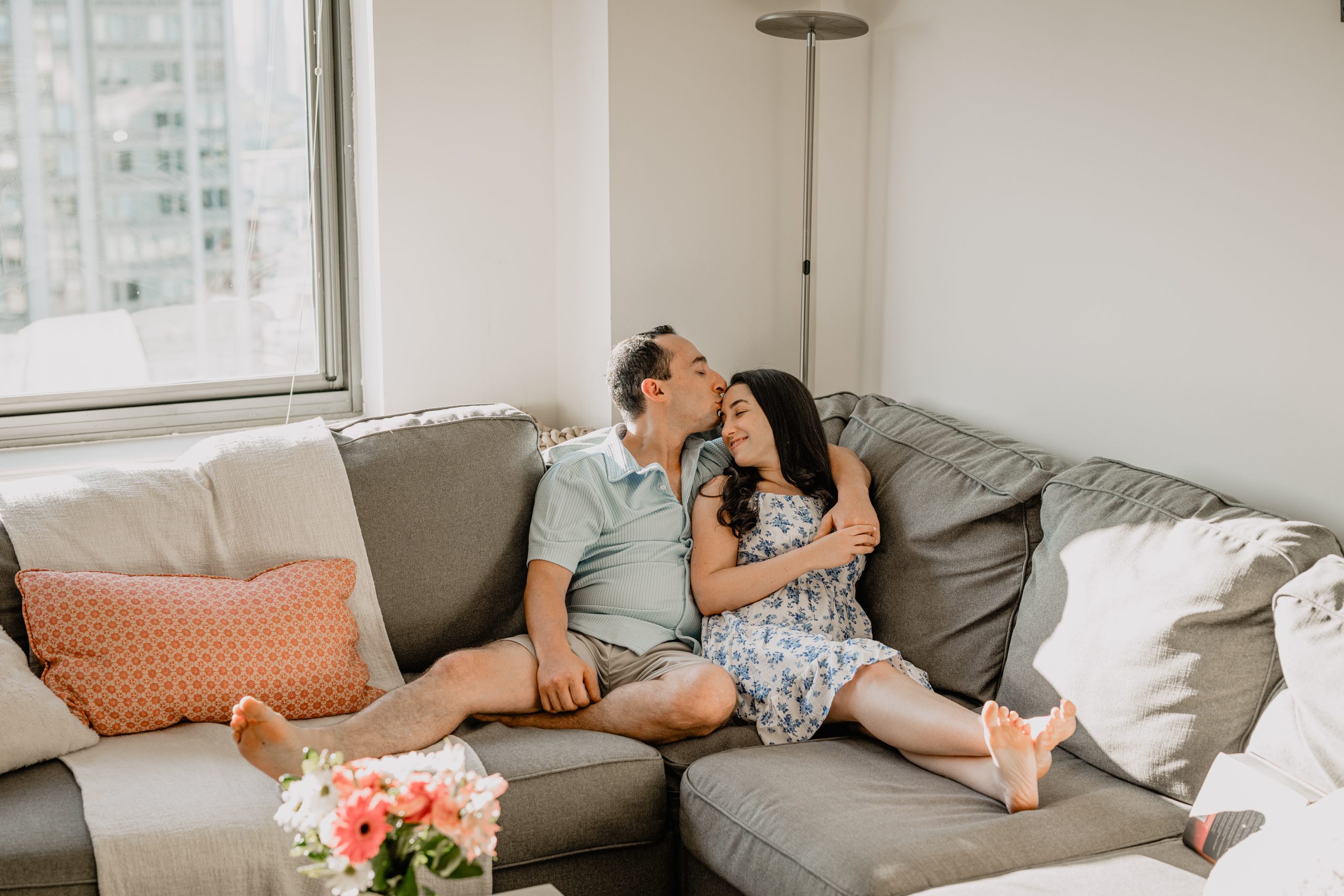 A sweet moment during a Jersey City & Home Engagement, with the couple sitting on their couch as the man gently kisses the woman on the forehead.