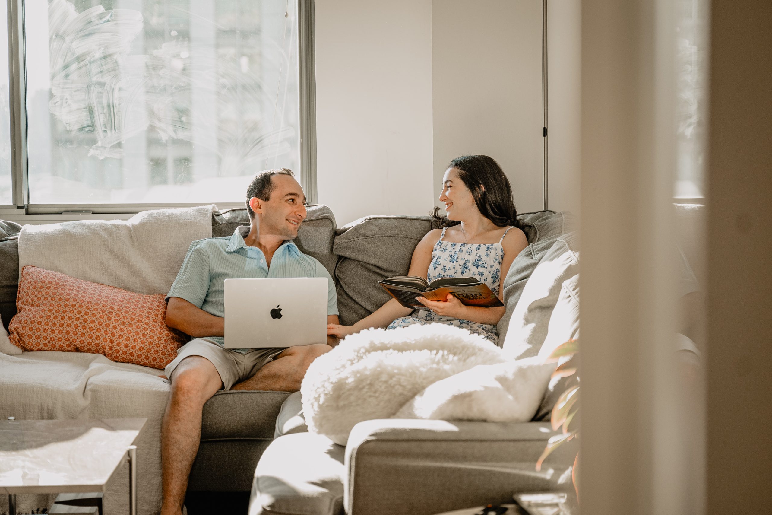 A cozy Jersey City & Home Engagement moment of the couple on their couch, with the man on his laptop and the woman reading a book as they look at each other.