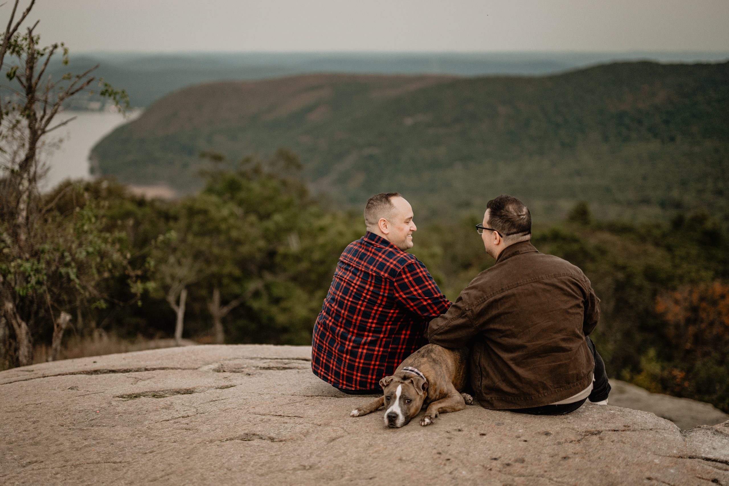Trevor and Josh sitting on the rocks with Duke resting beside them at their Bear Mountain engagement.