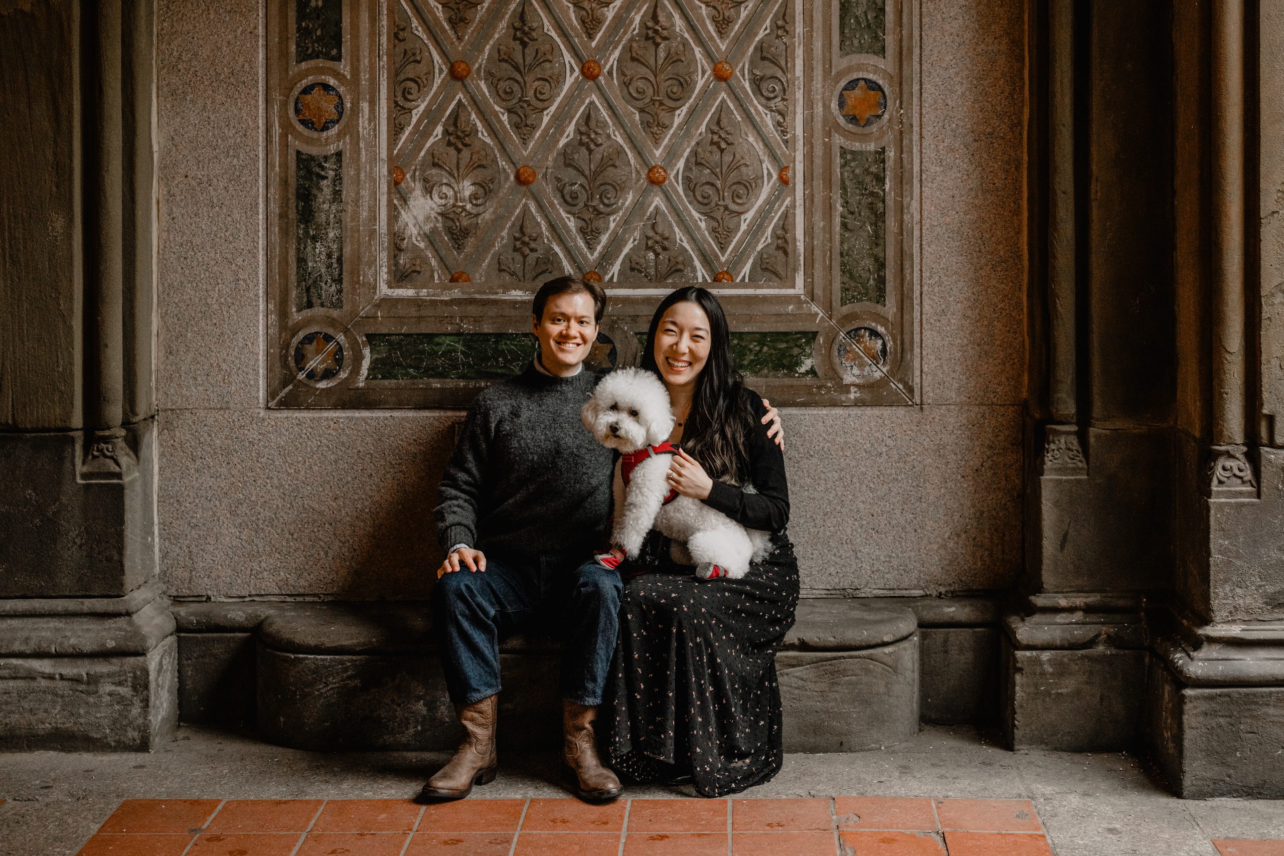 Kimberly and Peter smiling together during their fall engagement session at Central Park, holding their dog Mochi under an archway.