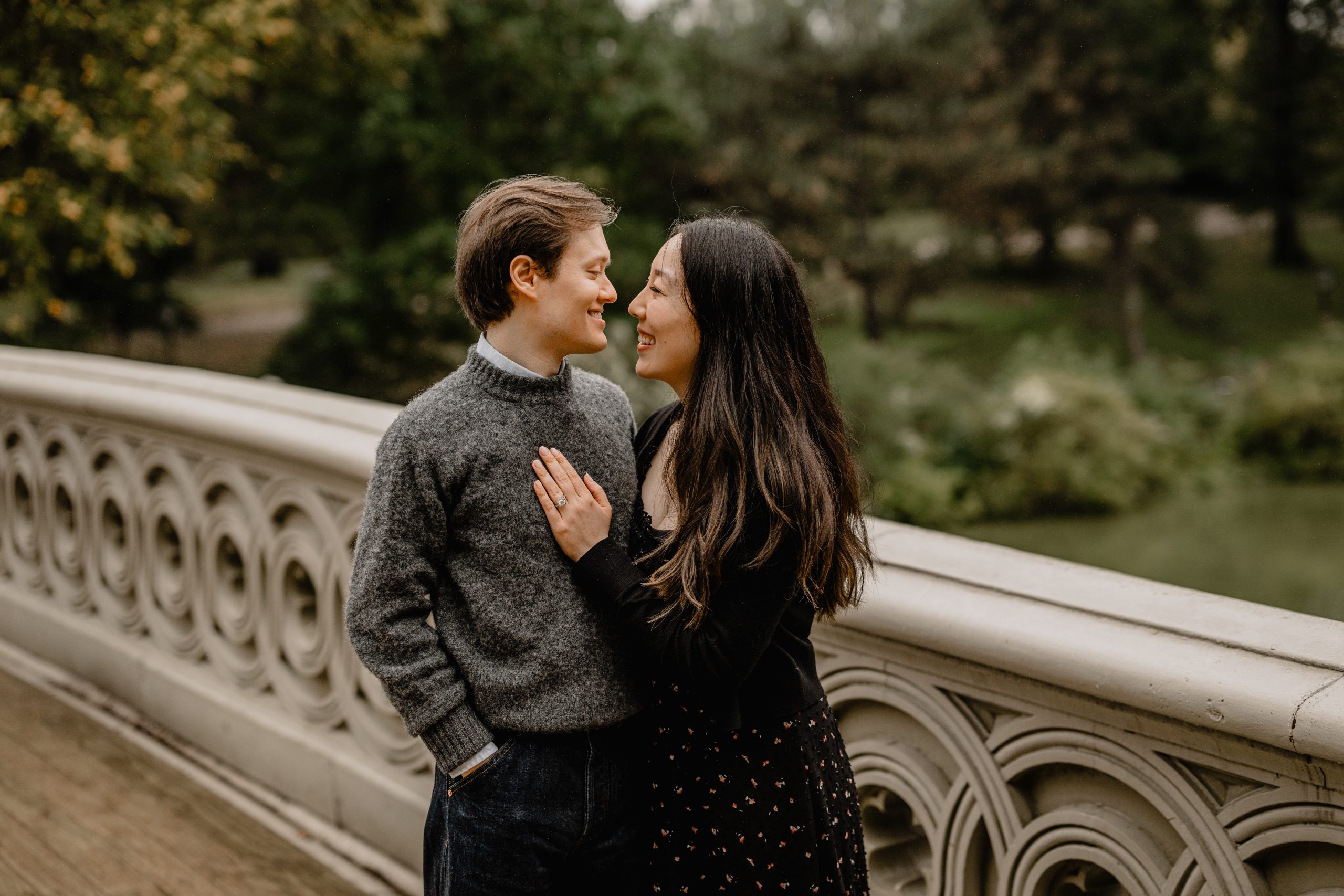 Kimberly and Peter standing on the Bow Bridge during their fall engagement session at Central Park, smiling at each other.
