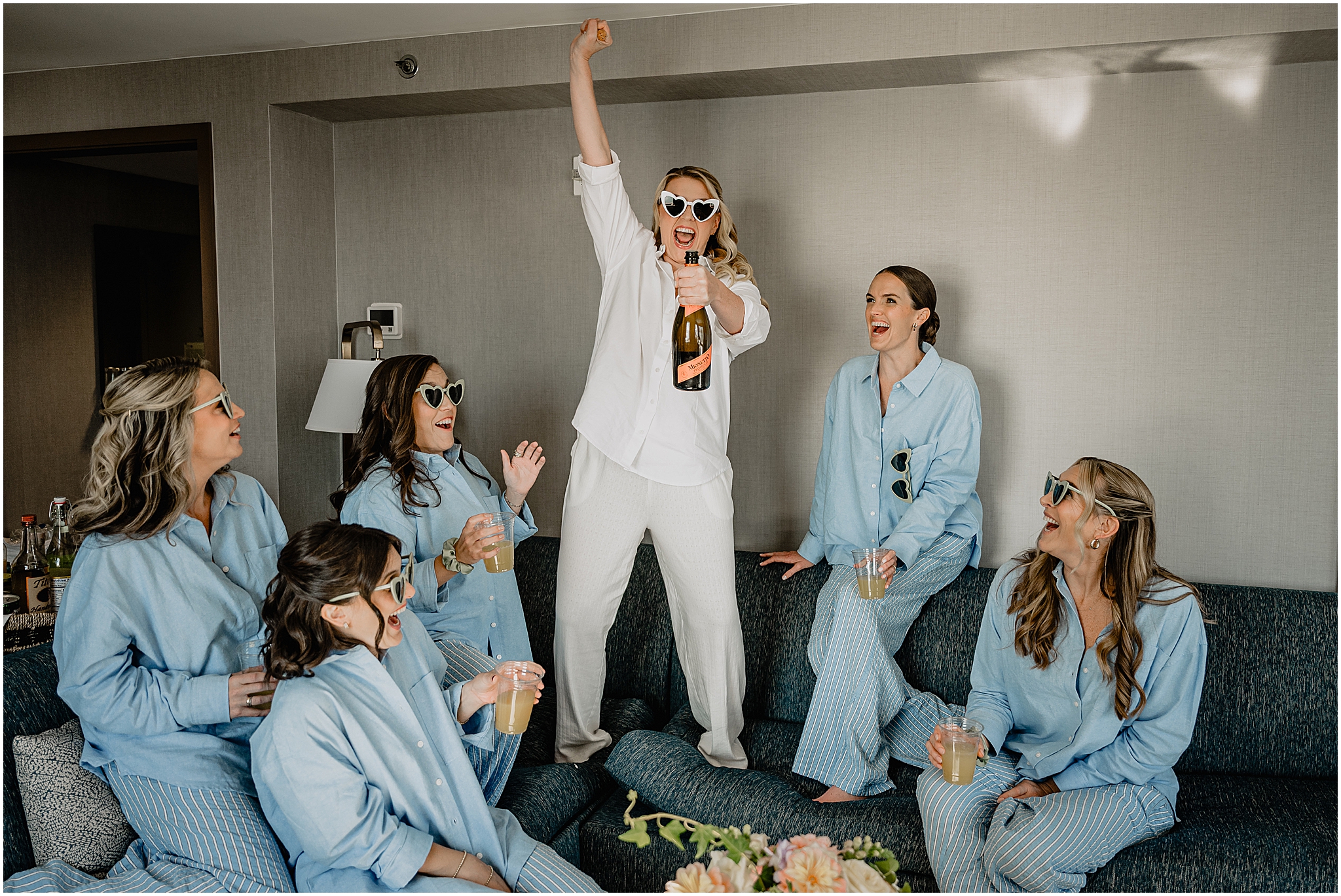 Bride holding up a bottle of wine while getting ready in pajamas, with the bridesmaids wearing heart-shaped sunglasses before their Colorful Fall Wedding at Liberty House.