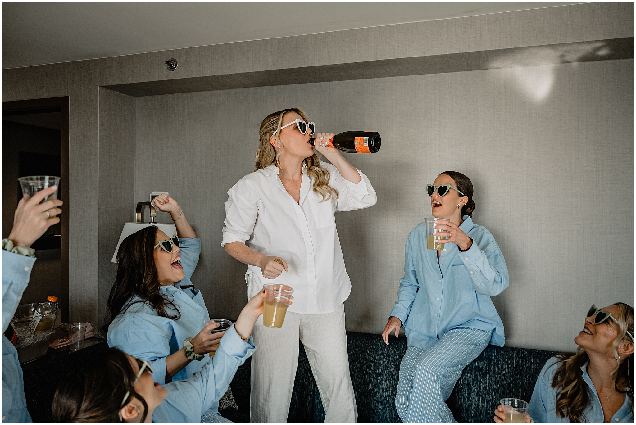 Bride drinking a bottle of wine while getting ready in pajamas, with the bridesmaids wearing heart-shaped sunglasses before their Colorful Fall Wedding at Liberty House.